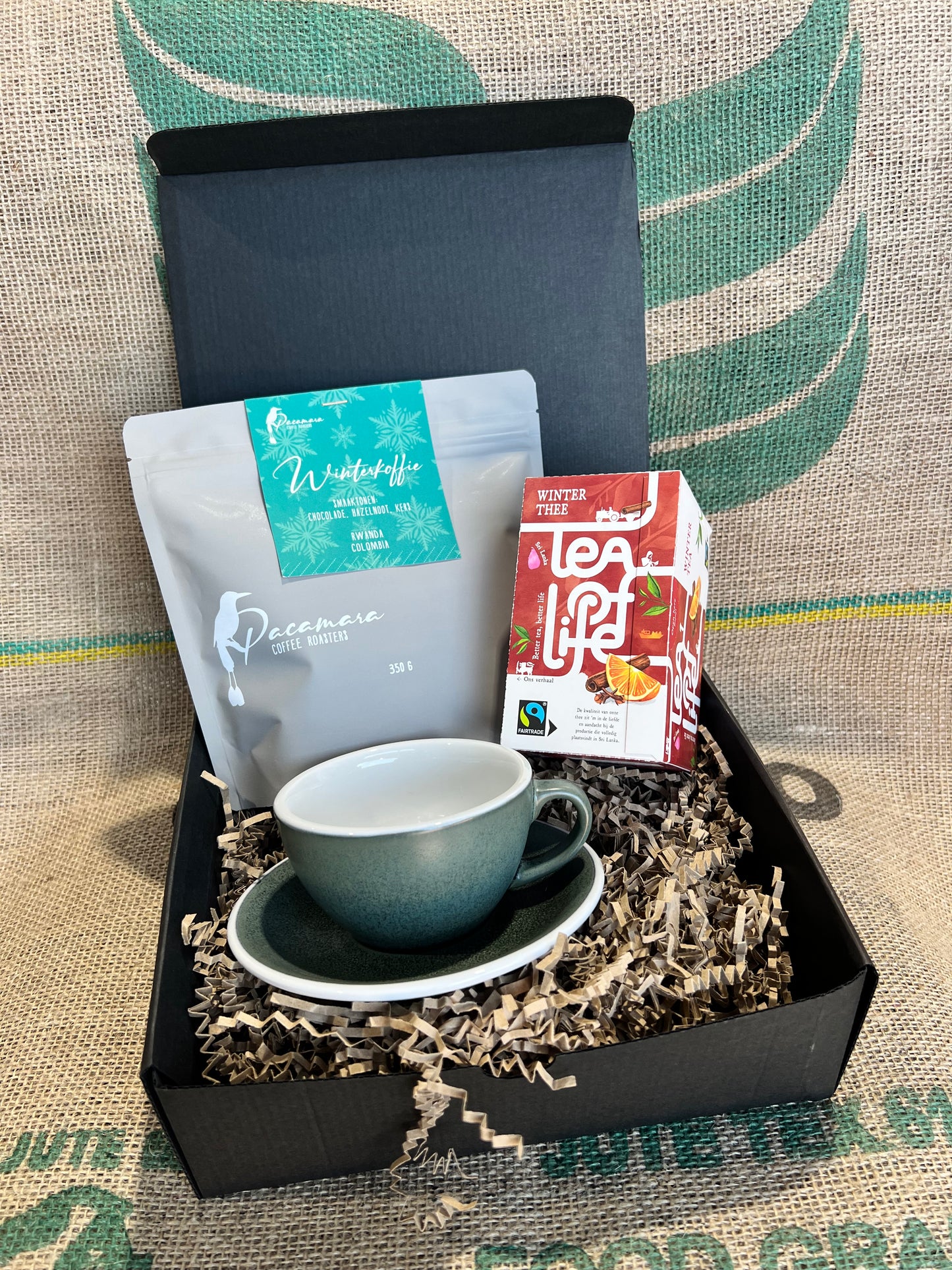 Zwarte cadeauverpakking, gevuld met papiersnippers. Inhoud bestaat uit een zakje winterkoffie, een doosje winterthee en een groene kop en schotel.